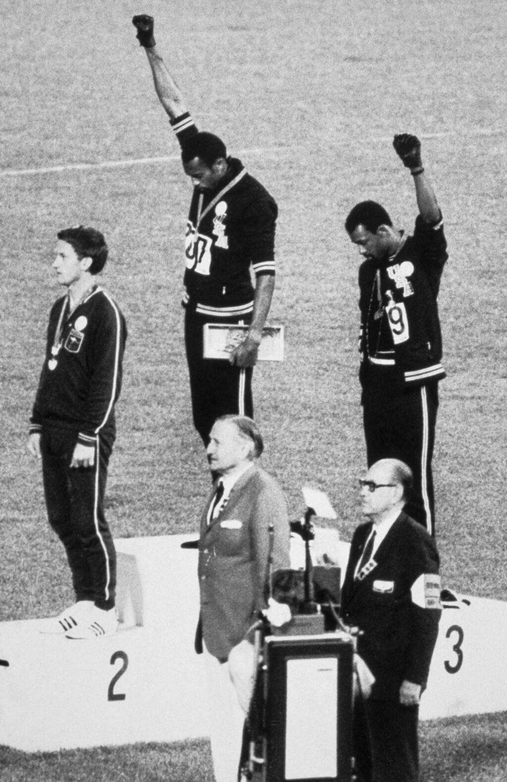 Les athlètes américains Tommie Smith (au centre), médaillé d'or, et John Carlos (à droite), médaillé de bronze, lèvent chacun un poing fermé et inclinent la tête pendant l'hymne national américain en signe de protestation pour les droits de l'homme lors de la remise des prix de la finale du 200 mètres masculin aux Jeux olympiques d'été à Mexico (Mexique) le 16 octobre 1968.