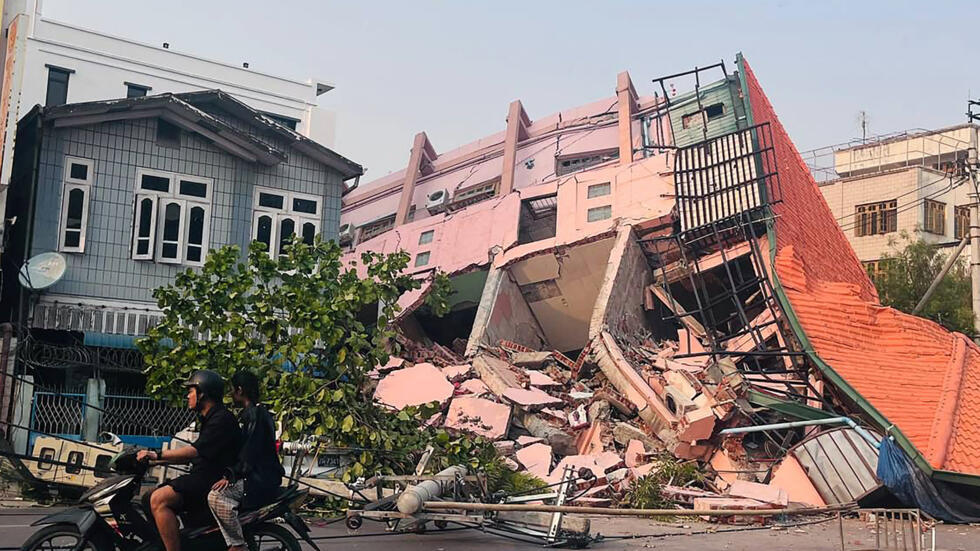 Des personnes passent en moto devant un bâtiment effondré à Mandalay, le 28 mars 2025, après un tremblement de terre.