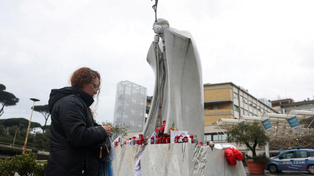 Mulher reza pelo papa Francisco diante da estátua de João Paulo 2°, instalada na entrada do hospital Gemelli, em Roma.