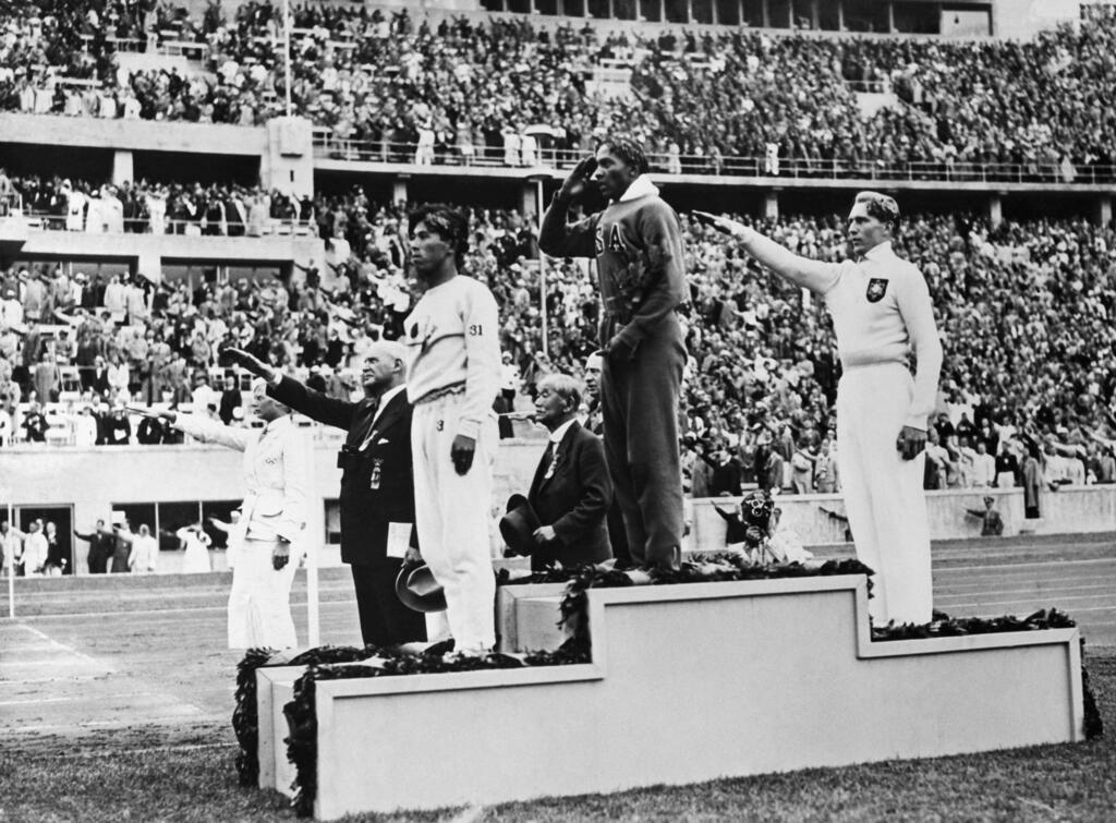Les médaillés d'or, d'argent et de bronze du concours olympique de saut en longueur se saluent depuis la tribune de la victoire aux Jeux olympiques d'été de 1936 à Berlin, le 8 août 1936. De gauche à droite, le Japonais Naoto Tajima (bronze), l'Américain Jesse Owens (or), qui a établi un record olympique dans cette épreuve et fait un salut à l'américaine en portant la main à son front, et l'Allemand Luz Long (argent), qui fait un salut nazi en tendant le bras.