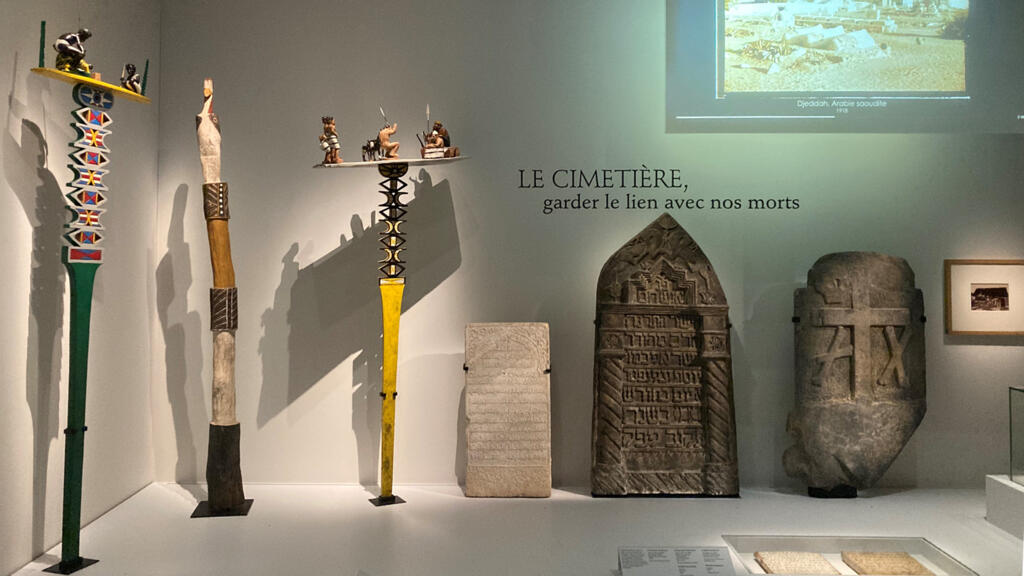 Dans l’exposition « Mourir, quelle histoire ! » sur les rites funéraires au Musée de Bretagne à Rennes : ici à droite des pierres tombales musulmane, juive et chrétienne et à gauche, deux poteaux funéraires de Madagascar (aloalo) en bois sculpté et peint et un poteau cérémoniel Pukamani d’Australie.