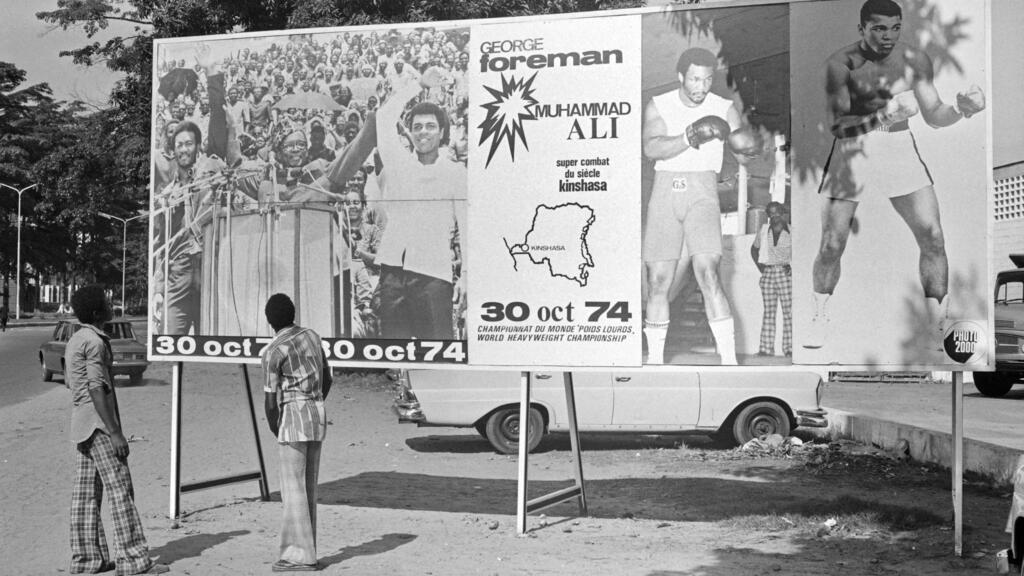 Le 30 octobre 1974, à Kinshasa, des gens regardent un panneau annonçant le combat entre les champions américains de boxe poids lourds, Mohamed Ali et George Foreman.