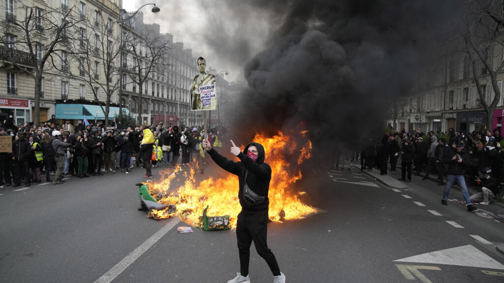 Un manifestant tient une pancarte affichant Emmanuel Macron dans le cortège parisien contre la réforme des retraites, le 28 mars 2023, aux côtés de poubelles en feu.