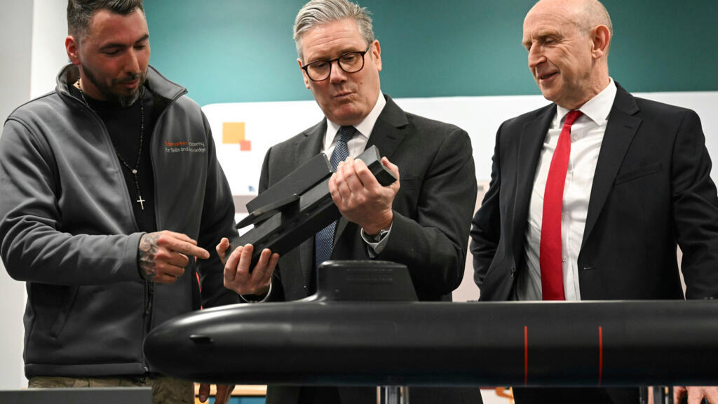 O primeiro-ministro britânico Keir Starmer, ao centro, e o secretário de Defesa John Healey, à direita, se reúnem com aprendizes do sistema BAE enquanto observam um modelo de submarino durante uma visita conjunta em Barrow-in-Furness, Inglaterra, na quinta-feira, 20 de março de 2025.