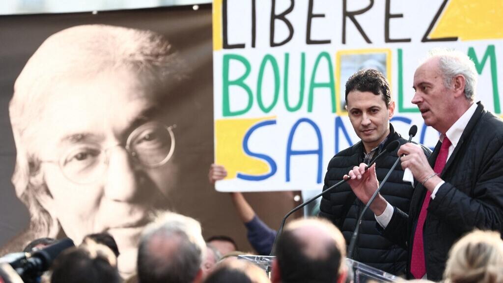 Le dessinateur de presse algérien Ghilas Aïnouche (2e à gauche) regarde le secrétaire général du comité de soutien Arnaud Benedetti prononcer un discours lors d'un rassemblement de soutien à l'écrivain franco-algérien détenu Boualem Sansal, à l'initiative de son comité de soutien, à côté d'une banderole à l'effigie de l'écrivain (à gauche), à Paris le 25 mars 2025. (Image d'illustration)