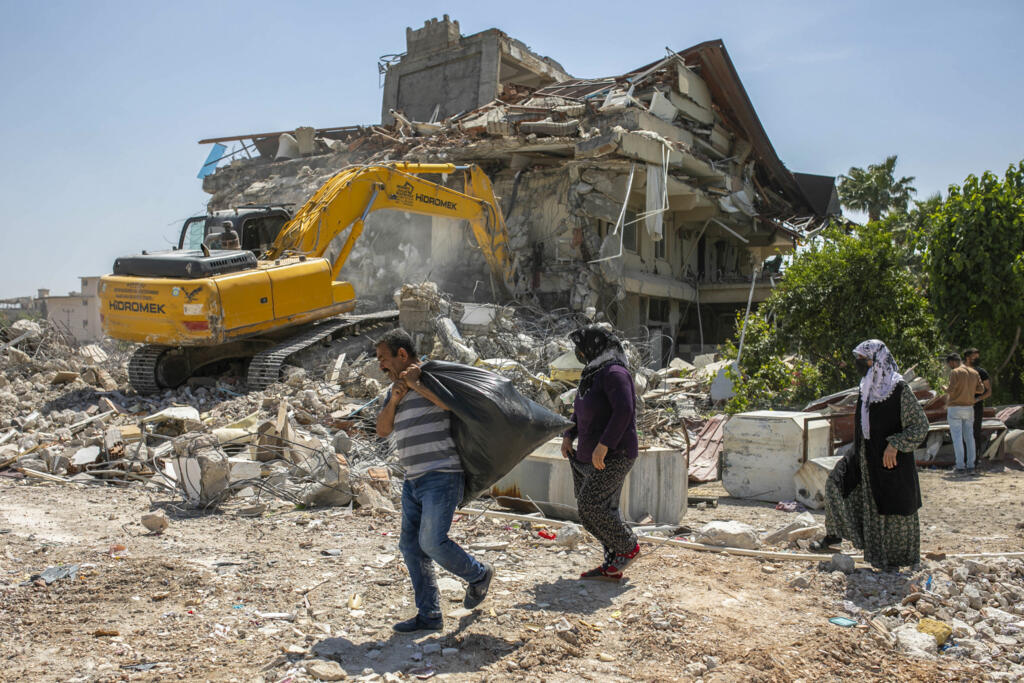 People walk past an excavator demolishing the remains of a destroyed building after a powerful earthquake in Antakya, southeastern Turkey, Wednesday, May 10, 2023.